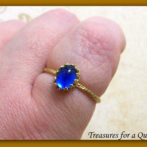 Puede incluir: Un anillo de oro con una piedra preciosa ovalada azul engastada en un engaste de oro. El anillo tiene una banda texturizada.