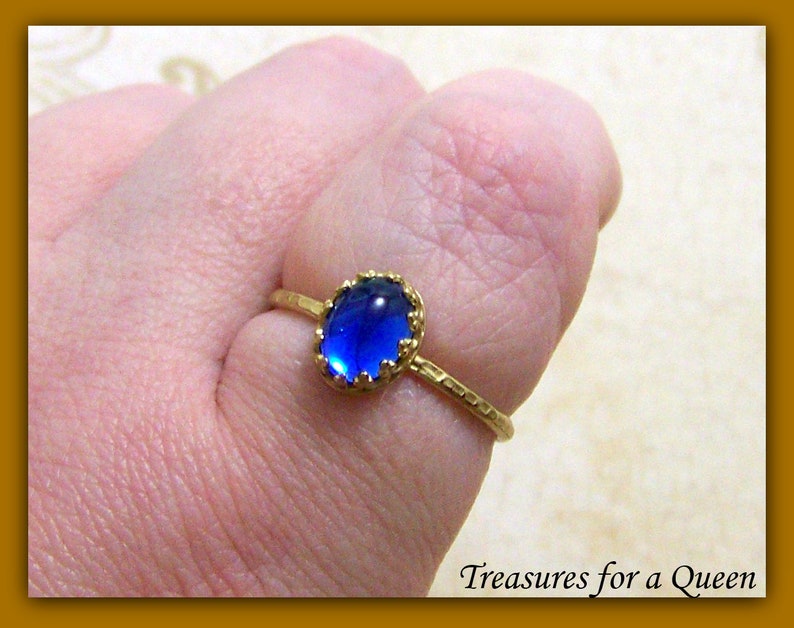 Puede incluir: Un anillo de oro con una piedra preciosa ovalada azul engastada en un engaste de oro. El anillo tiene una banda texturizada.