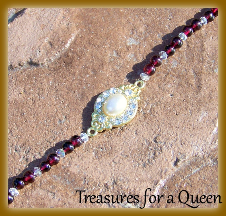 May include: A gold-tone necklace with a large, oval-shaped pendant featuring a white pearl surrounded by clear rhinestones. The necklace is made of small, dark red and clear beads.