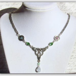 Puede incluir: Un collar de plata con un nudo celta adornado con cuentas de cristal verde y transparente. El colgante presenta un dise&ntilde;o de nudo detallado con una sola gota de cristal transparente.