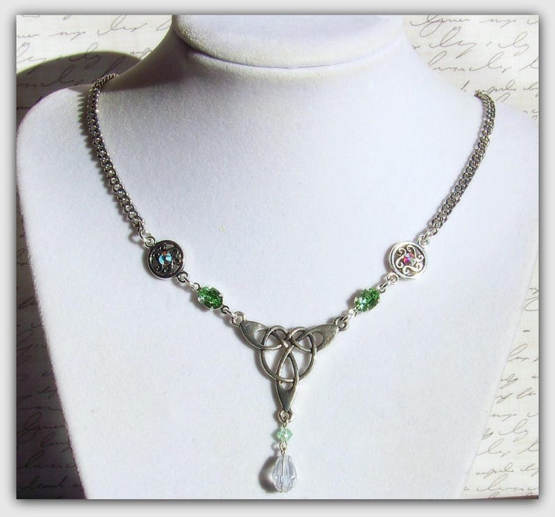 Puede incluir: Un collar de plata con un nudo celta adornado con cuentas de cristal verde y transparente. El colgante presenta un dise&ntilde;o de nudo detallado con una sola gota de cristal transparente.