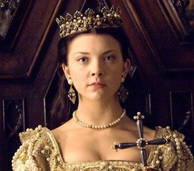 May include: A woman wearing a gold crown with red and white gems, a pearl necklace, and pearl earrings. She is holding a silver cross with pearl accents. She is wearing a gold and white dress.