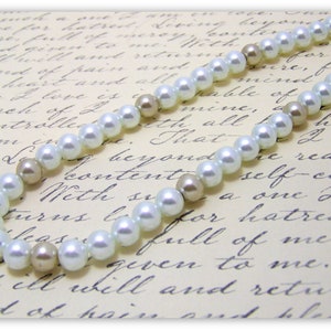 May include: A white pearl necklace with beige pearl accents. The necklace is on a white background with black cursive writing.