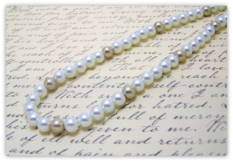 May include: A white pearl necklace with beige pearl accents. The necklace is on a white background with black cursive writing.