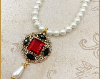 May include: A gold necklace with a red and black pendant. The pendant is a round, ornate design with a large red square gem in the center and black oval gems around it. The necklace is made of white pearls.