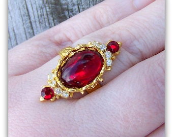 May include: A gold ring with a large red oval gemstone surrounded by smaller clear gemstones. The ring is set in a gold filigree design.