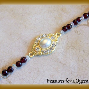 May include: A gold-tone bracelet with a large oval-shaped pendant featuring a faux pearl surrounded by clear rhinestones. The bracelet is made of small dark red beads and clear beads.