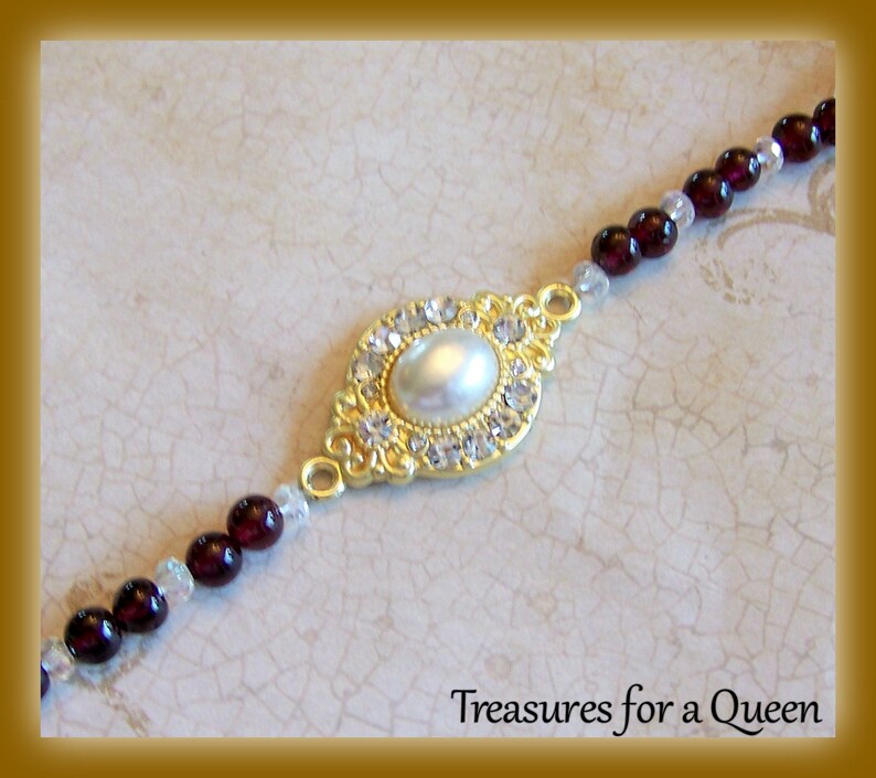 May include: A gold-tone bracelet with a large oval-shaped pendant featuring a faux pearl surrounded by clear rhinestones. The bracelet is made of small dark red beads and clear beads.