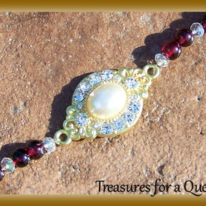 May include: A gold-tone bracelet with a large, oval-shaped pendant featuring a white pearl surrounded by clear rhinestones. The bracelet is made of small, dark red beads and clear rhinestones.
