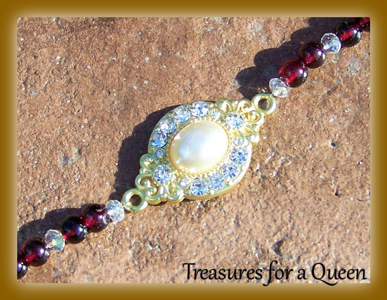 May include: A gold-tone bracelet with a large, oval-shaped pendant featuring a white pearl surrounded by clear rhinestones. The bracelet is made of small, dark red beads and clear rhinestones.