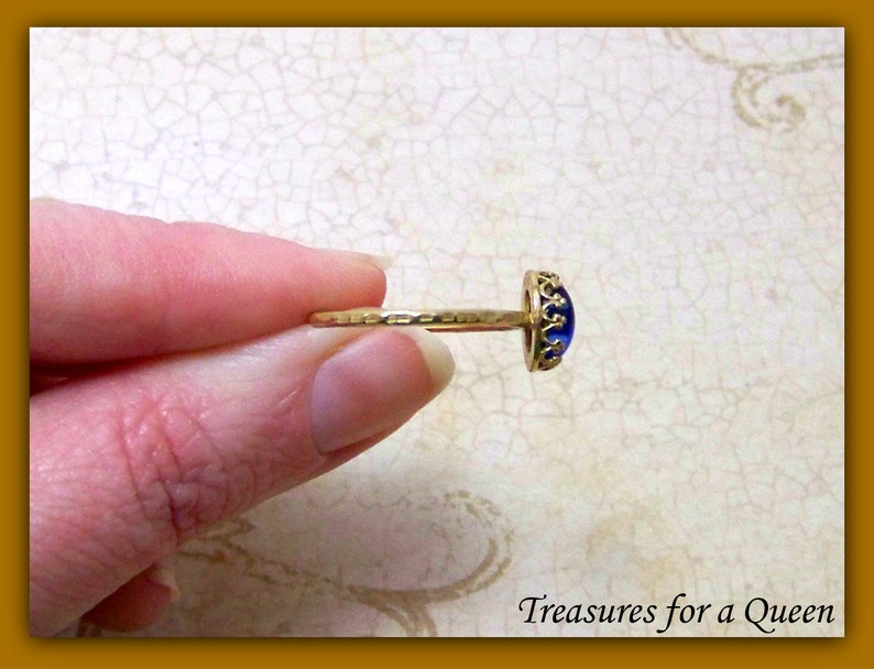 Puede incluir: Un anillo de oro con una piedra de vidrio azul engastada en un bisel con forma de corona. El anillo est&aacute; en un dedo.