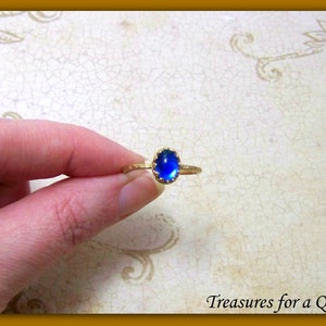 Puede incluir: Un anillo de oro con una piedra preciosa ovalada azul engastada en un engaste en forma de corona. El anillo est&aacute; en un dedo.
