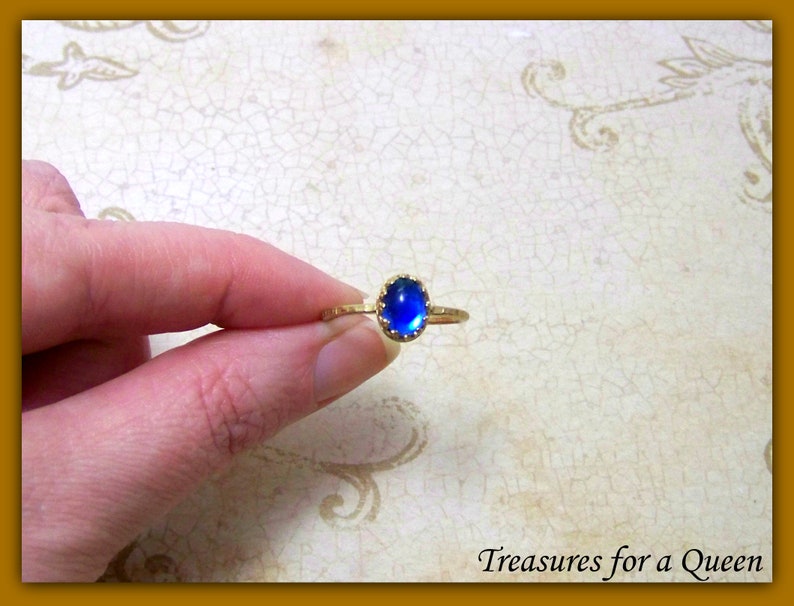 Puede incluir: Un anillo de oro con una piedra preciosa ovalada azul engastada en un engaste en forma de corona. El anillo est&aacute; en un dedo.