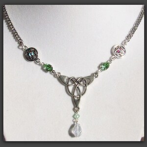 Puede incluir: Un collar de plata con un nudo celta, acentos de cristal verde y azul. El collar tiene un colgante de gota con un cristal transparente.