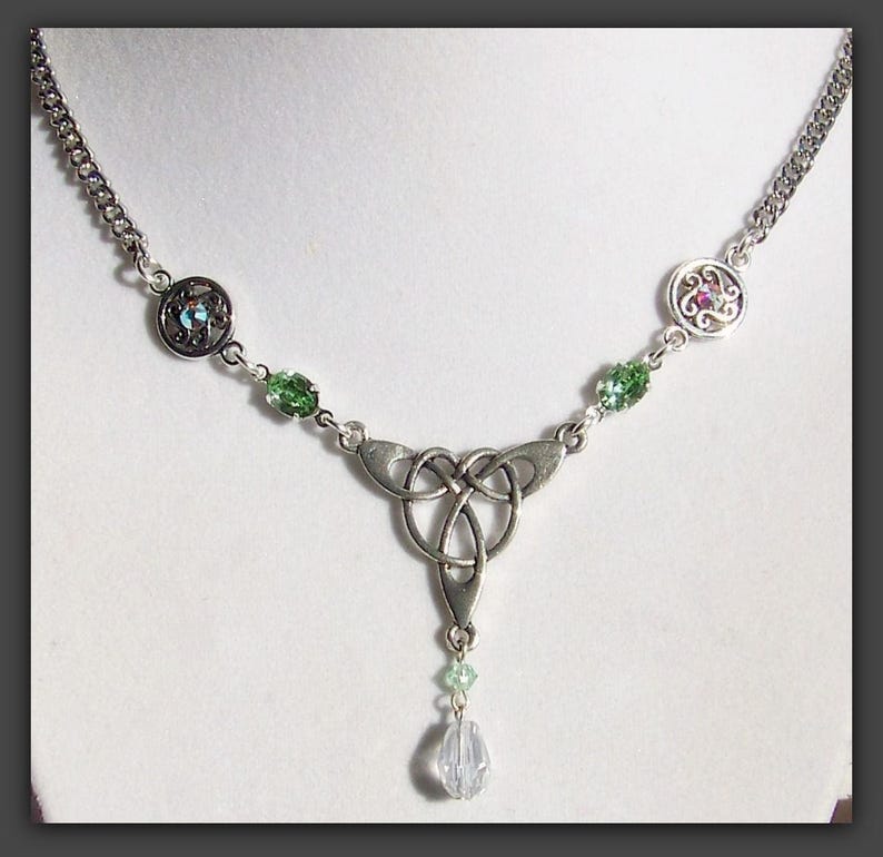Puede incluir: Un collar de plata con un nudo celta, acentos de cristal verde y azul. El collar tiene un colgante de gota con un cristal transparente.