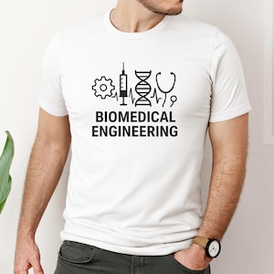 Puede incluir: Camiseta blanca con un diseño gráfico negro que presenta símbolos médicos y de ingeniería, incluyendo un engranaje, una jeringa, una cadena de ADN y un estetoscopio. Las palabras "BIOMEDICAL ENGINEERING" están impresas debajo.