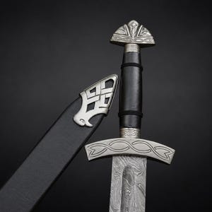 May include: A sword with a patterned Damascus steel blade, a black leather-wrapped grip, and a silver-toned metal pommel and crossguard featuring intricate engravings. Beside the sword is a black leather scabbard with a silver metal chape.