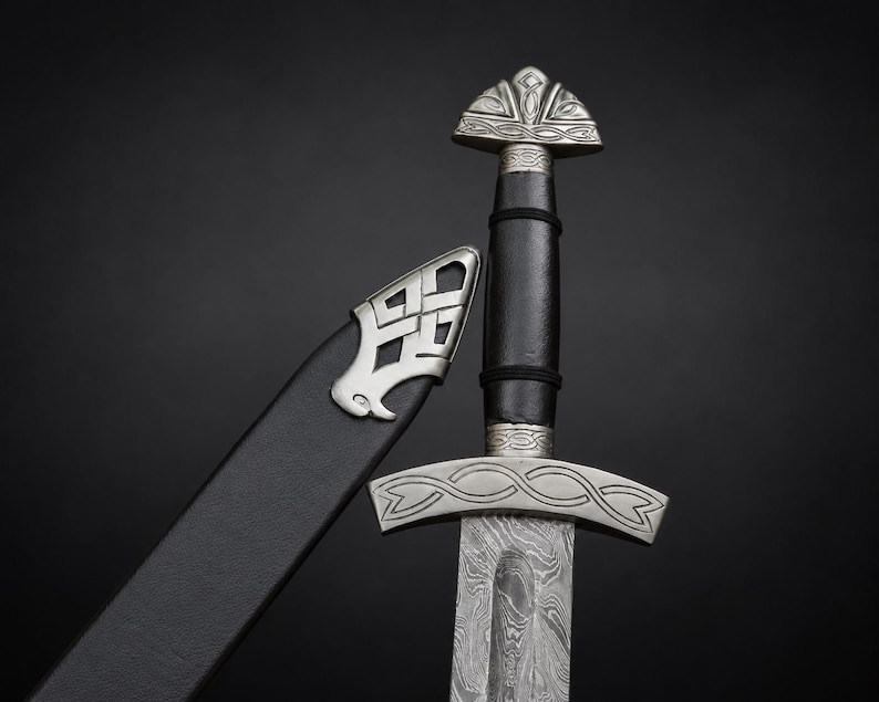 May include: A sword with a patterned Damascus steel blade, a black leather-wrapped grip, and a silver-toned metal pommel and crossguard featuring intricate engravings. Beside the sword is a black leather scabbard with a silver metal chape.