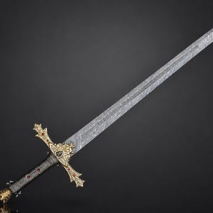 May include: A long sword featuring a silver-toned blade with a distinct wavy Damascus steel pattern. The hilt is crafted with ornate gold-colored metal accents, including a lion head motif, and is inlaid with small red stones along the crossguard.