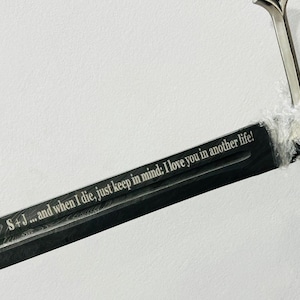 May include: A long sword with a dark, patterned blade featuring the engraved text: S + J ... and when I die, just keep in mind: I love you in another life! The sword has a silver-colored crossguard and a black handle wrapped in clear plastic film.
