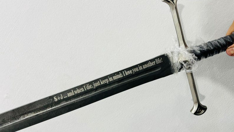 May include: A long sword with a dark, patterned blade featuring the engraved text: S + J ... and when I die, just keep in mind: I love you in another life! The sword has a silver-colored crossguard and a black handle wrapped in clear plastic film.