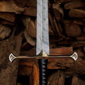 May include: A sword with a double-edged Damascus steel blade featuring a wavy pattern, a polished brass crossguard with triangular gold-colored inlays at the ends, and a black leather-wrapped handle with a central ridge running down the blade.