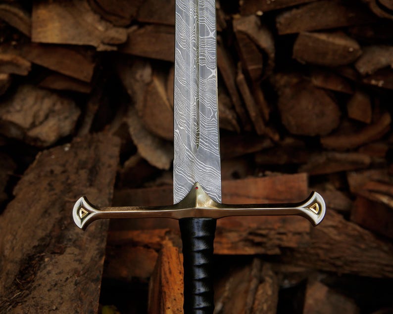 May include: A sword with a double-edged Damascus steel blade featuring a wavy pattern, a polished brass crossguard with triangular gold-colored inlays at the ends, and a black leather-wrapped handle with a central ridge running down the blade.