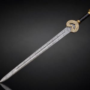 May include: A straight sword with a double-edged blade featuring a visible folded steel pattern. The hilt includes a gold-colored metal guard with ornate scrollwork, a black cylindrical grip, and a matching gold-colored pommel at the end.