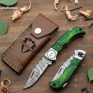 May include: Two folding knives with green handles and a brown leather sheath. The knives have silver blades with a unique pattern. The sheath is embossed with a tuxedo design and the text "GROOMSMAN - 20-05-2025".