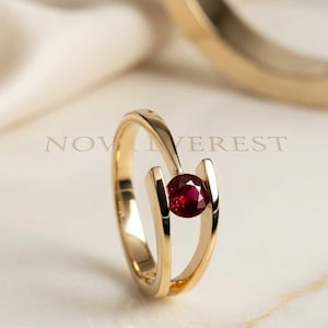 May include: A gold ring featuring a round, deep red gemstone set between two open, polished gold bands. The text NOV EVEREST is visible in the background. The design showcases a tension-style setting that holds the vibrant red stone securely.