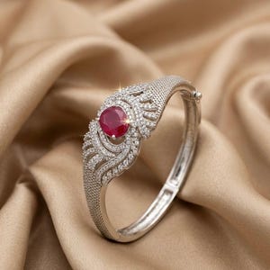 Könnte beinhalten: Silberfarbenes Armband mit einem großen, runden, roten Edelstein in der Mitte. Das Armband ist mit kleinen, klaren Edelsteinen und aufwendigen Filigranmustern verziert. Der Hintergrund ist ein drapiertes, goldbraunes Gewebe.
