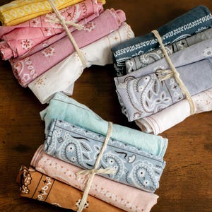 May include: Bundles of rolled bandanas in various colors, including yellow, pink, blue, and brown. Each bundle is tied with twine. The bandanas feature paisley and other decorative patterns.