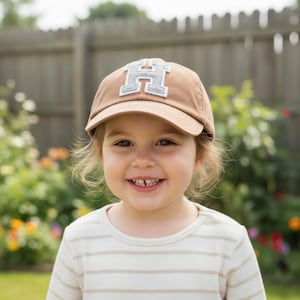 May include: A tan baseball cap featuring a large, centered letter H in a light gray, textured fabric with a white border. The cap has a curved brim and a structured crown, constructed from a solid-colored cotton material with visible stitching.