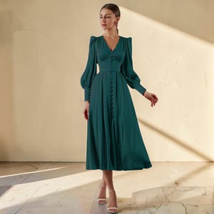 Puede incluir: Un vestido largo de color verde oscuro con escote en V y mangas largas. El vestido presenta una botonadura frontal y una cintura entallada. Las mangas son abullonadas en los hombros y se estrechan en las mu&ntilde;ecas. El vestido se combina con tacones blancos.