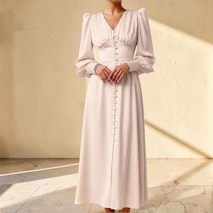 Puede incluir: Vestido largo rosa p&aacute;lido con escote en V y mangas largas. El vestido presenta una fila de botones en la parte delantera y en los pu&ntilde;os. El vestido tiene un corpi&ntilde;o ajustado y una falda fluida, ideal para ocasiones especiales.