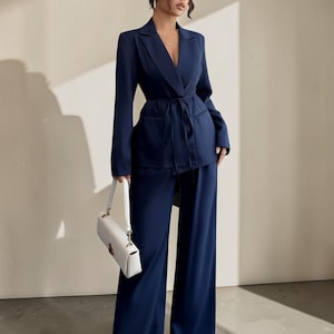 Puede incluir: Un conjunto de traje azul marino con una blazer con cinturón anudado y pantalones de pernera ancha. La blazer tiene mangas largas y solapas con muescas. Se sostiene un bolso blanco con asa superior.