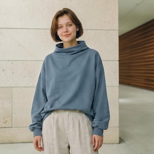 Peut inclure: Un sweat-shirt à manches longues bleu poussiéreux doté d'un col montant et d'une coupe ample et décontractée. Le vêtement est associé à un pantalon beige clair présentant un détail plissé visible sur le devant au niveau de la taille.