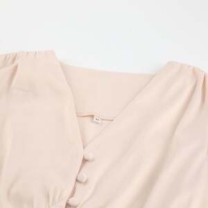 Puede incluir: Blusa de color melocot&oacute;n claro con cuello en V y cierre de botones. La tela parece ligera. El dise&ntilde;o es sencillo y elegante, adecuado para diversas ocasiones. La blusa tiene una etiqueta con la letra 'M'.