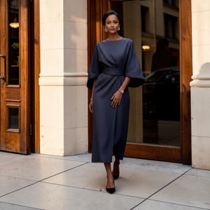 May include: A dark slate blue midi dress featuring a wide round neckline, loose elbow-length sleeves, and a fitted waist band. The skirt has a straight silhouette with a subtle wrap-style front detail. The garment is paired with black pointed heels.