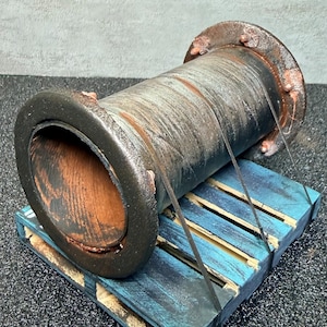 May include: A weathered, cylindrical metal pipe with rust-colored accents rests on a small, blue-painted wooden pallet. The pipe's interior is visible, showing a dark, textured surface. The pallet has a distressed appearance.