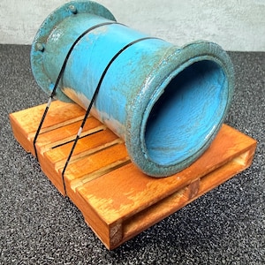 May include: A large, cylindrical, turquoise-colored pipe with a weathered appearance, secured with black straps, rests on a small wooden pallet. The pallet has a natural wood finish. The background is a dark, textured surface.
