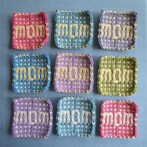 Puede incluir: Nueve salvamanteles tejidos de colores con la palabra "MOM" en el centro. Los salvamanteles son de varios colores, incluyendo rosa, azul, morado y verde, con la palabra "MOM" en un color crema contrastante.