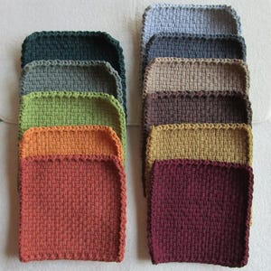 Puede incluir: Una colección de posavasos cuadrados tejidos en varios colores, incluyendo tonos de verde, naranja y burdeos. Los posavasos tienen un aspecto texturizado y un borde cosido. La disposición muestra los diferentes tonos y texturas.