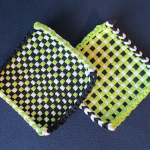 Könnte beinhalten: Zwei quadratische, gewebte Topflappen mit einem Schachbrettmuster in Schwarz, Weiß und Limettengrün. Die Kanten sind mit einem kontrastierenden schwarz-weißen Zopf verziert. Die Topflappen liegen auf einer dunkelblauen Oberfläche.