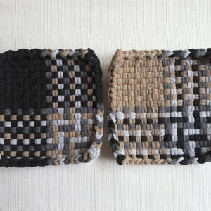 May include: Two square potholders featuring a woven, checkered pattern in shades of black, tan, gray, and white. The potholders are constructed from thick, braided fabric strips with a visible, textured grid design and a braided border edge.