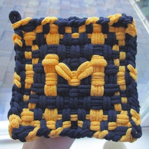 Op de afbeelding: Geweven vierkante pannenlap in marineblauw en goud. De pannenlap heeft een grote gouden letter "M" in het midden. De randen zijn afgewerkt met een lus om op te hangen. Het geweven ontwerp is zichtbaar over de hele pannenlap.