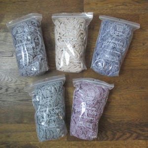Peut inclure: Cinq sachets en plastique transparent contenant des boucles en coton pour le tissage, étiquetés Friendly Loom traditional size cotton loops makes 6 potholders. Les sachets contiennent des boucles grises, beiges, bleu-gris et violettes.