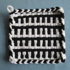 Puede incluir: Salvamanteles cuadrado tejido en blanco y negro con un lazo para colgar. El diseño se asemeja a las teclas de un piano. Los bordes están acabados con un borde trenzado. El salvamanteles mide unos 18 cm cuadrados.