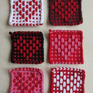 Pode incluir: Uma coleção de seis pegadores quadrados, tecidos em várias combinações de cores. Os pegadores apresentam tons de vermelho, branco, preto e rosa, com um padrão geométrico texturizado. Cada pegador tem uma pequena alça para pendurar.