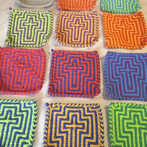 Op de afbeelding: Een verzameling kleurrijke, geweven pannenlappen. Elke vierkante pannenlap heeft een kruisontwerp in verschillende kleurencombinaties, waaronder rood en oranje, blauw en paars, en groen en geel. De pannenlappen hebben een gevlochten rand en een kleine lus om op te hangen.